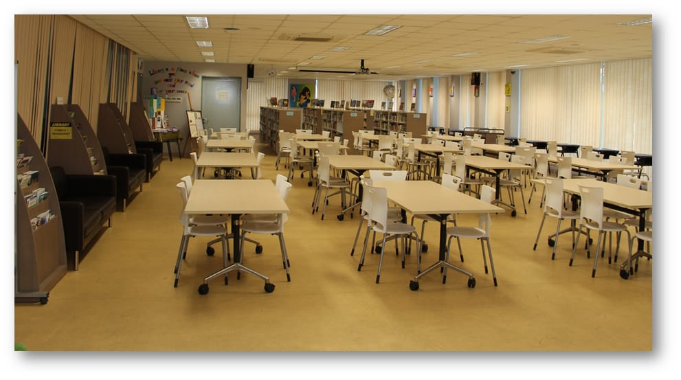 A spacious empty school library with white tables on wheels, white chairs, bookshelves, and vertical blinds on windows.